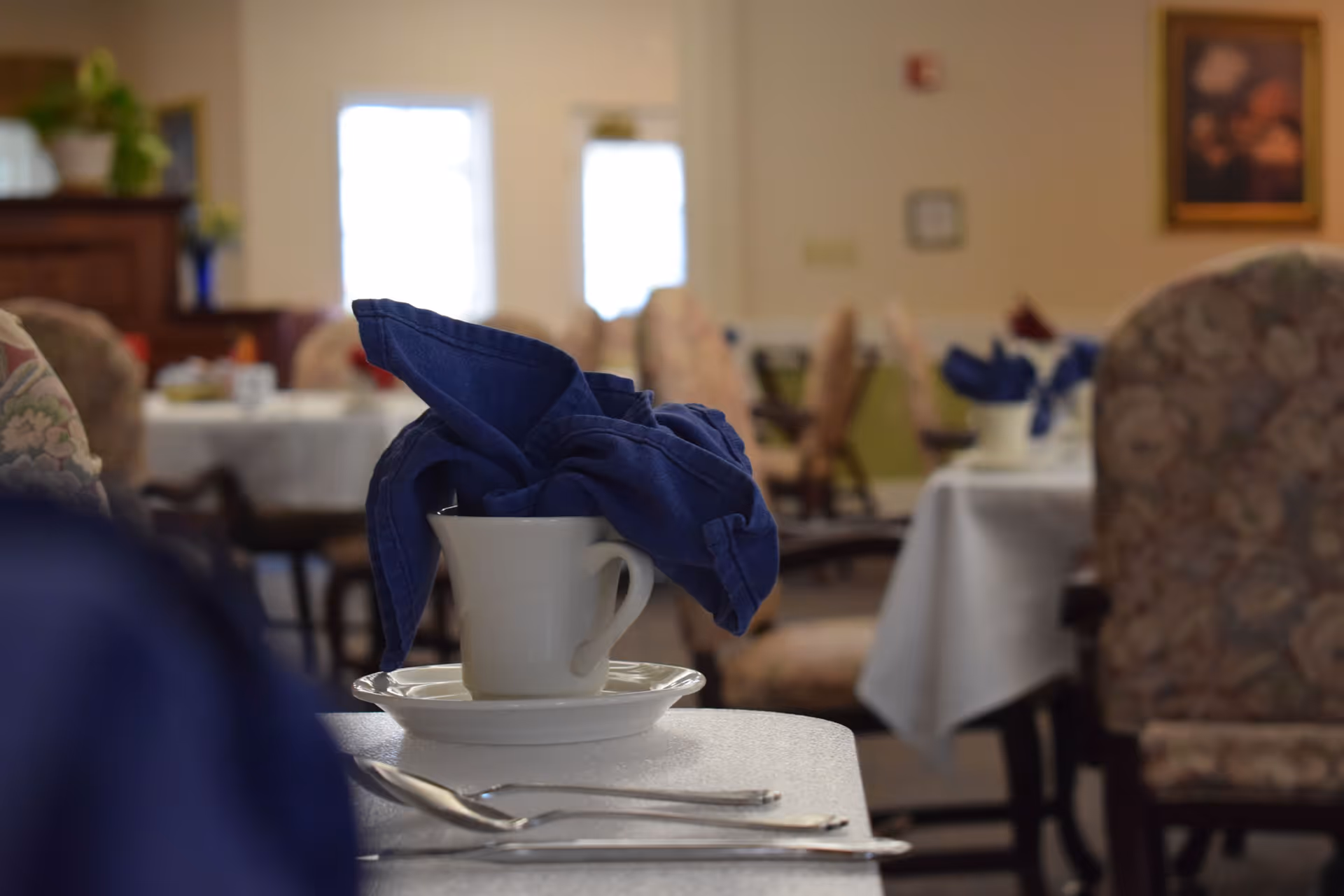A dining room table set with a teacup holding a folded blue cloth napkin, utensils in the foreground and blurred chairs and tables in the background.