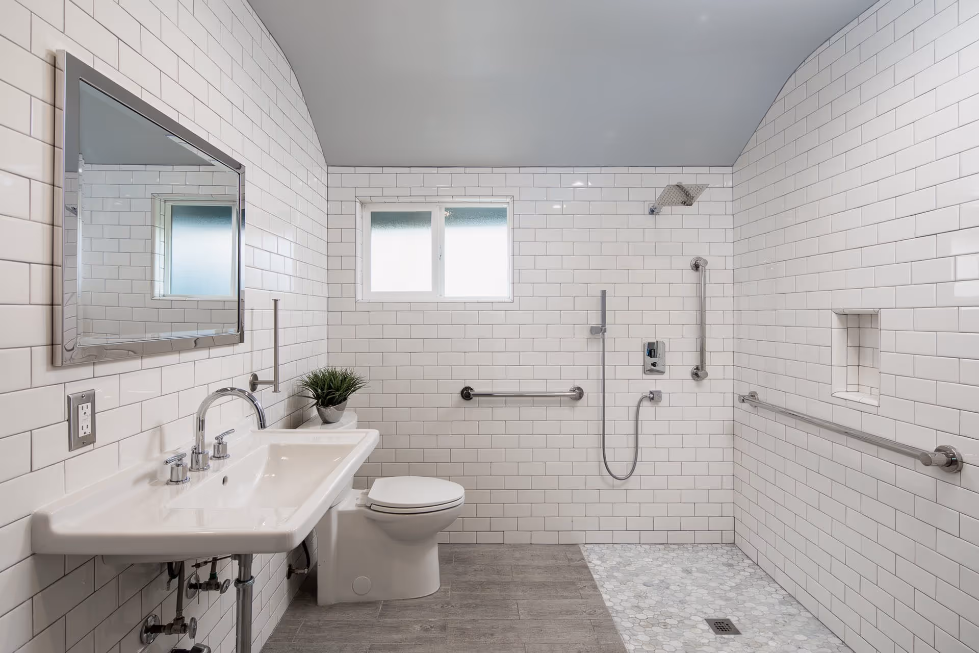 Bright accessible bathroom with white subway tile, wall-mounted sink and mirror, toilet, and a roll-in shower area with grab bars and handheld showerhead.