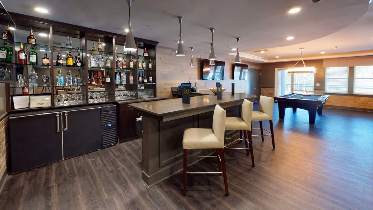 A modern indoor bar area with a long counter and three cushioned bar stools. Behind the bar, shelves are stocked with various bottles of liquor and glassware. The room features wood flooring, pendant lights hanging above the bar, two wall-mounted flat-screen TVs, and a pool table near large windows letting in natural light.