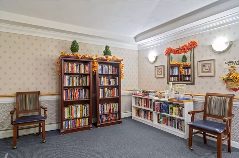 A cozy reading area in a senior living facility with two wooden bookshelves filled with books, decorated with autumn leaves and small green topiary plants on top. There are two wooden chairs with cushioned seats on either side of the bookshelves. A white built-in bookshelf below a mirror also holds books and a water pitcher with cups. The walls are decorated with patterned wallpaper, framed pictures, and wall sconces providing soft lighting.