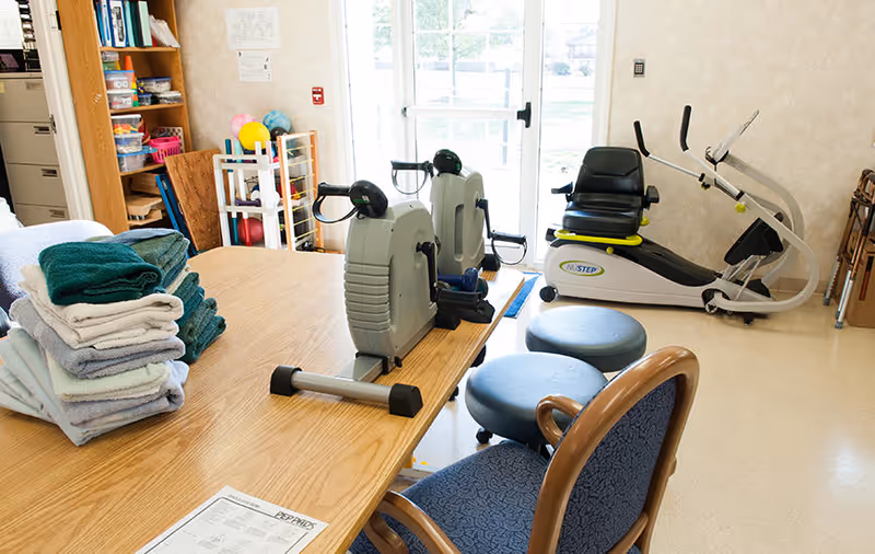 A room with exercise equipment including two stationary pedal exercisers on a wooden table and a recumbent exercise bike near a glass door. There are folded towels stacked on the table, two blue cushioned stools, a blue chair, and shelves with various items in the background.
