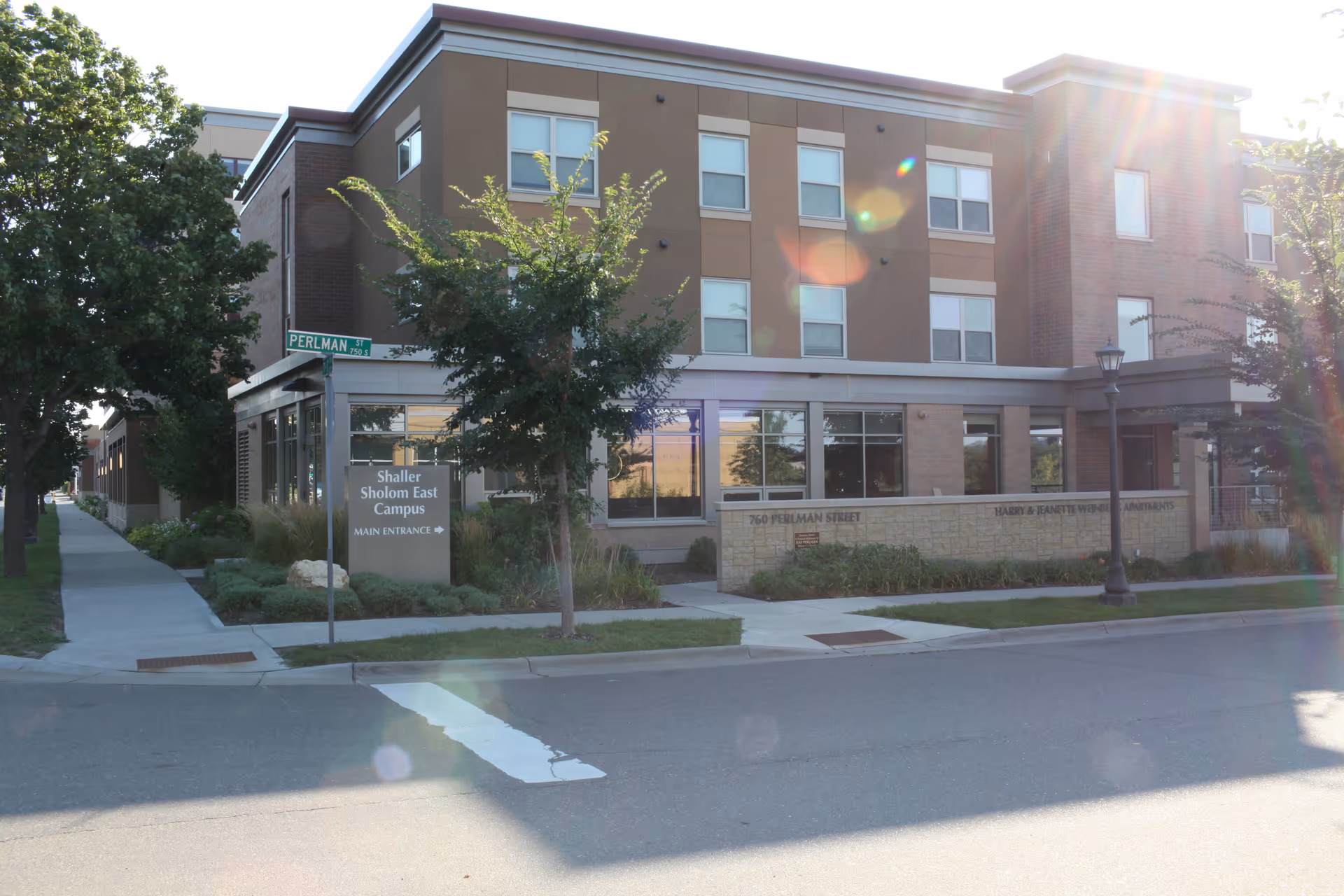 Exterior view of the Shaller Sholom East Campus building on Perlman Street, showing a modern three-story structure with large windows, trees, and a sidewalk in front. A sign near the entrance reads 'Shaller Sholom East Campus MAIN ENTRANCE'.
