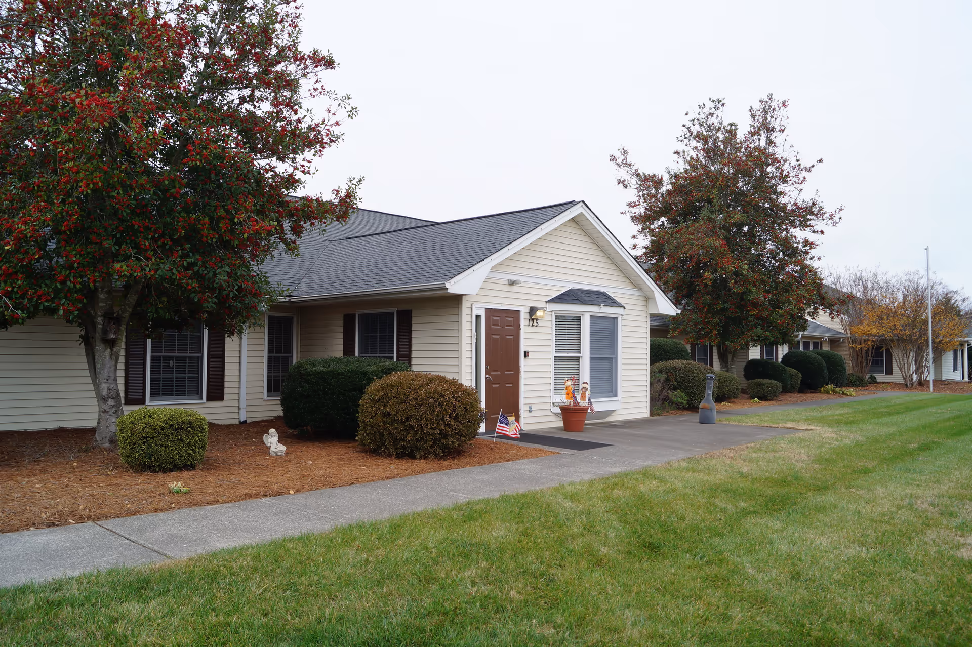 Exterior view of a single-story senior living facility building with beige siding and a dark shingled roof. The entrance door is brown with the number 125 above it. There are two windows with white blinds next to the door. The building is surrounded by neatly trimmed bushes and trees with red berries. A concrete walkway runs in front of the building, bordered by green grass and mulch beds with small decorative statues and potted plants near the entrance.