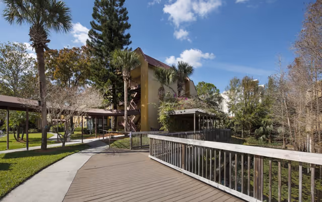 Outdoor view of Concordia Village of Tampa showing a wooden deck with railing over a pond, a paved walkway, palm trees, and a multi-story building with exterior staircases under a partly cloudy blue sky.