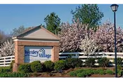 Outdoor view of the entrance sign for Wesley Village Senior Living, featuring a brick and stone sign with the facility name, surrounded by greenery, flowering trees, a white fence, and a street lamp.