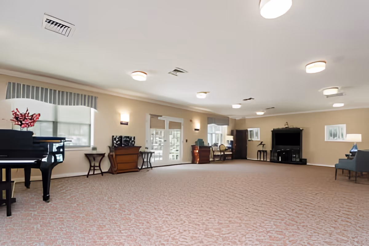 A spacious, well-lit common room with beige walls and carpeted floor. The room features a black grand piano with a vase of red flowers on top, several small tables and chairs, a fireplace, and large windows with valances allowing natural light to enter.