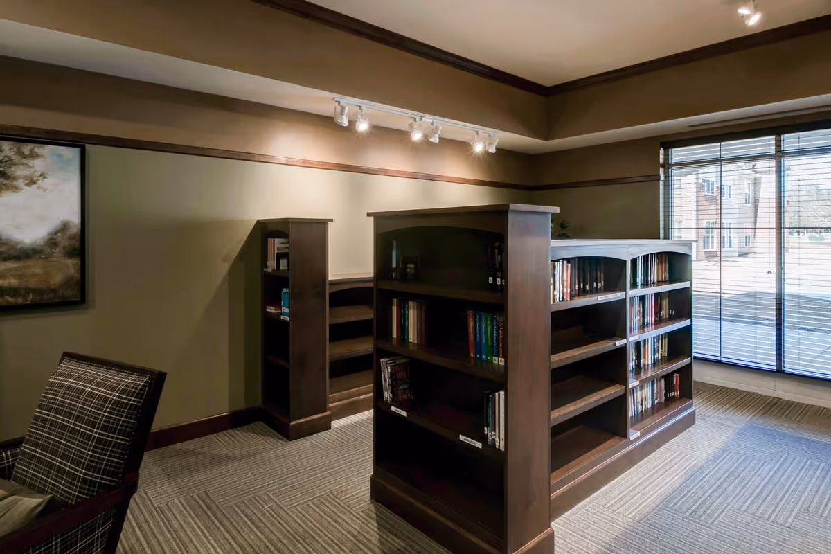 Interior view of a quiet library or reading area with wooden bookshelves filled with books, a plaid upholstered chair, carpeted floor, a large window with blinds letting in natural light, and a framed painting on the wall.