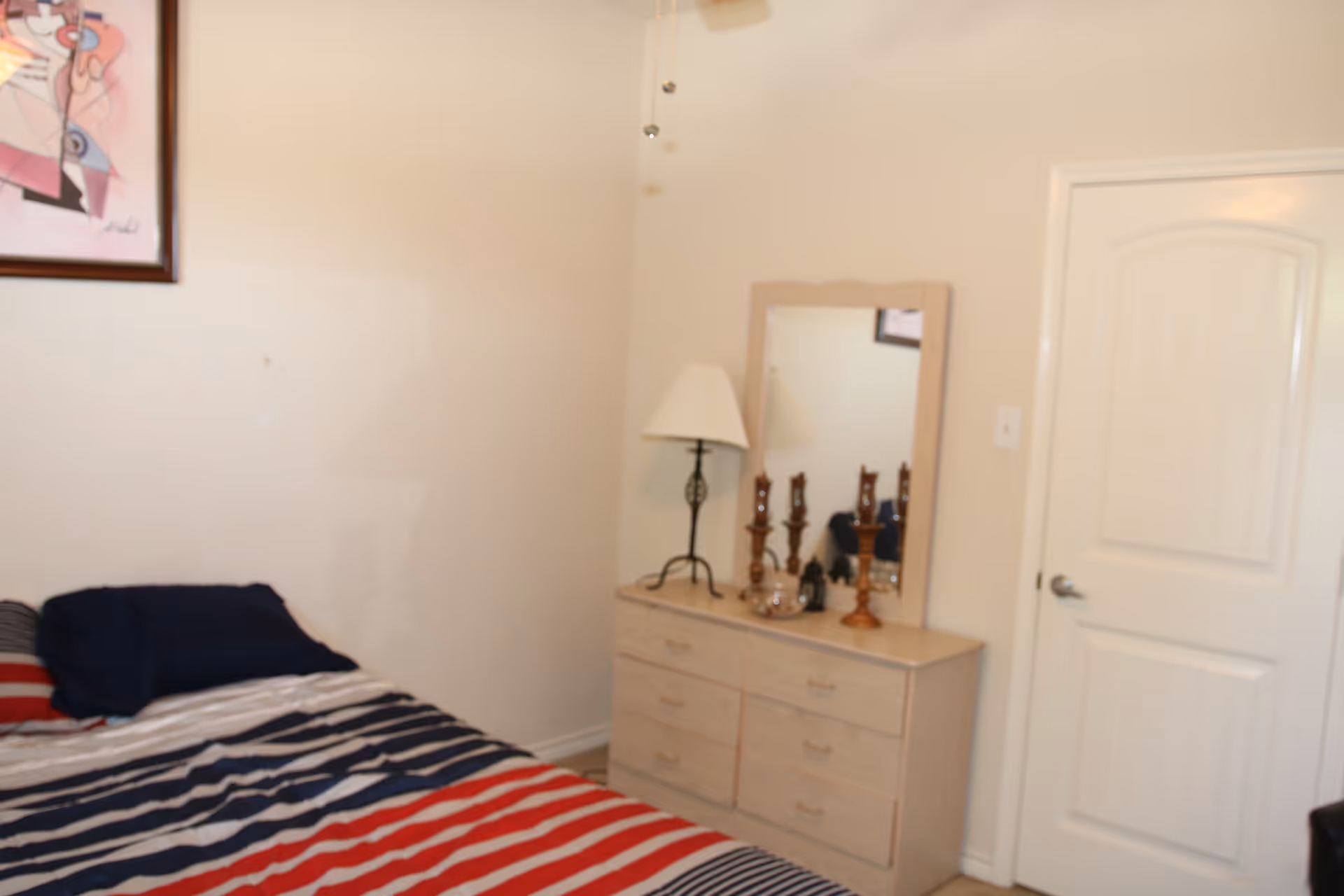 A bedroom with a bed covered in a striped red, white, and blue blanket, a dresser with a mirror, a table lamp, and decorative items. A closed white door is visible on the right side of the room, and a framed abstract artwork hangs on the wall above the bed.
