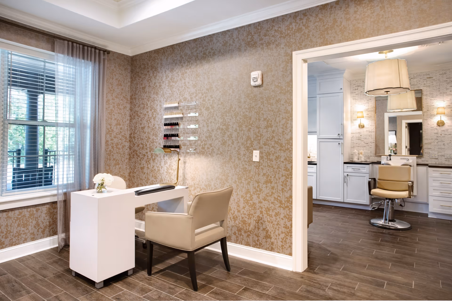 A well-lit beauty salon area in a senior living facility featuring a white manicure table with a beige armchair, a small vase with white flowers, and a wall-mounted display of nail polish. Through an open doorway, a hair styling station with a beige salon chair, large mirror, and white cabinetry is visible. The room has patterned beige wallpaper, wood-look tile flooring, and a window with sheer curtains.