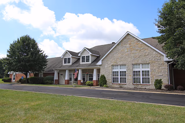 Exterior view of a single-story senior living facility building with a stone and brick facade, several windows, a covered entrance with white columns, an American flag, and a paved driveway. There are trees and green grass surrounding the building under a partly cloudy sky.