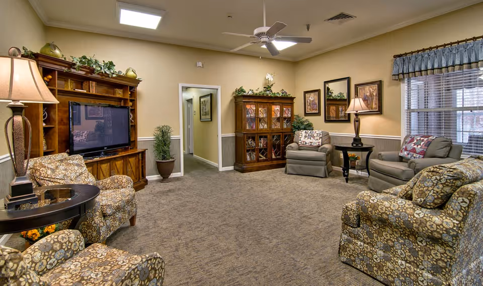 Cozy common living room with patterned armchairs and recliners arranged around a TV cabinet, lamps, and decorative shelving.
