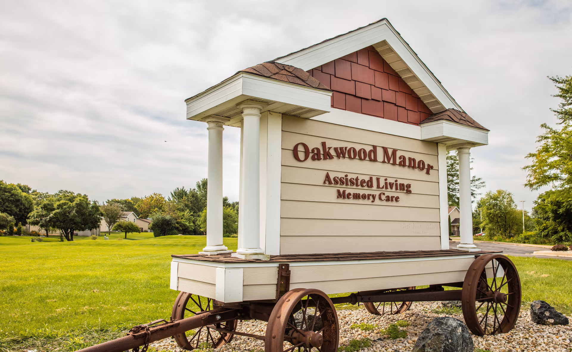 A large wooden sign for Oakwood Manor Assisted Living Memory Care, designed like a small house on wheels with white pillars and a shingled roof, situated on a grassy area with trees and houses in the background under a cloudy sky.