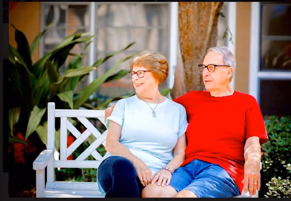 An elderly couple sitting closely together on a white wooden bench outdoors, smiling and enjoying each other's company. The woman is wearing a light blue shirt and glasses, and the man is wearing a red shirt and glasses. There are green plants and a tree in the background with a building window visible.