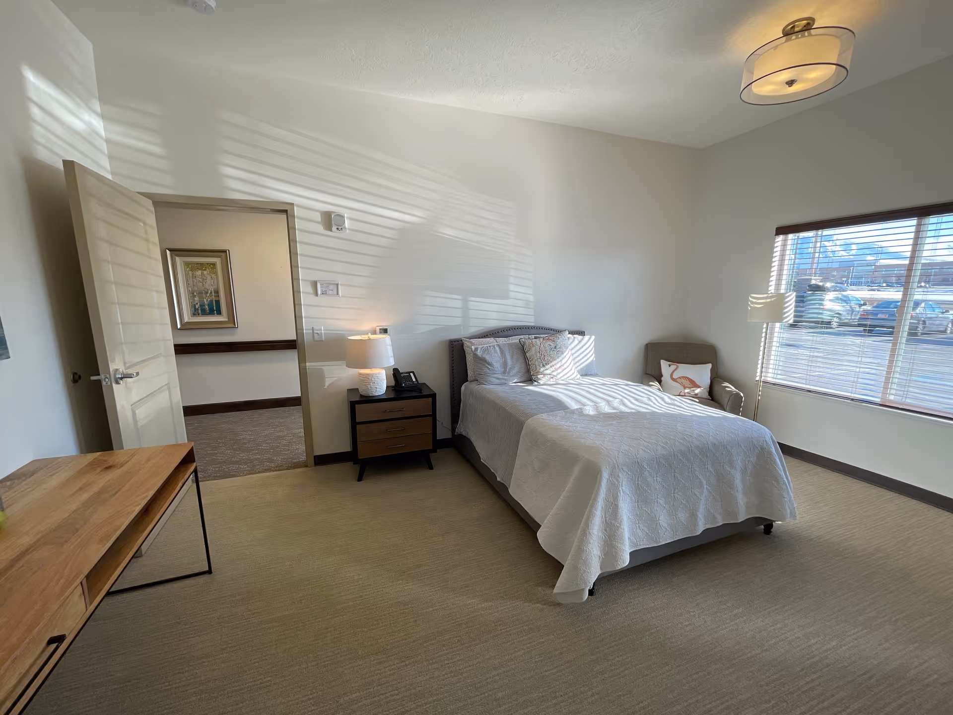 A bright and clean bedroom in an assisted living facility featuring a neatly made bed with white bedding, a bedside table with a lamp and phone, a comfortable armchair with a decorative pillow, a large window with blinds letting in natural light, and a wooden desk. The room has beige carpet and light-colored walls.