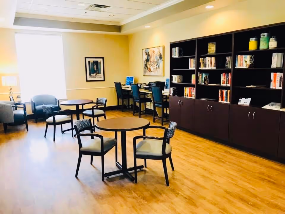 A well-lit room with wooden flooring featuring several round tables each surrounded by chairs. Along one wall is a large dark wood bookshelf filled with books and decorative items. In the corner, there is a desk area with computers and chairs. The room has light-colored walls with framed artwork and a window letting in natural light.