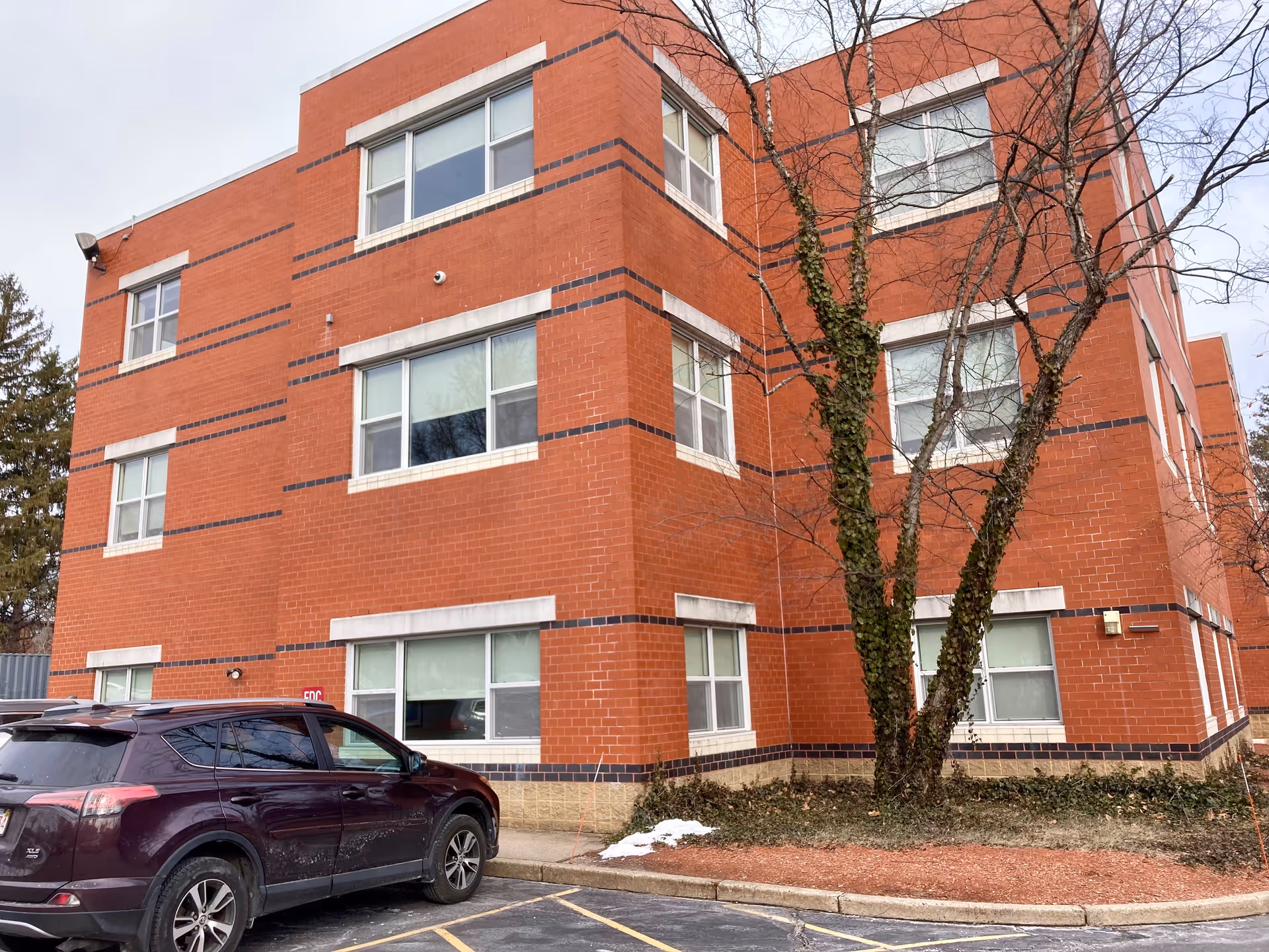 Exterior view of a three-story red brick building with multiple windows. A tree with bare branches and some ivy growing on its trunk is in front of the building. A dark-colored SUV is parked in a parking space near the building. The sky is overcast.
