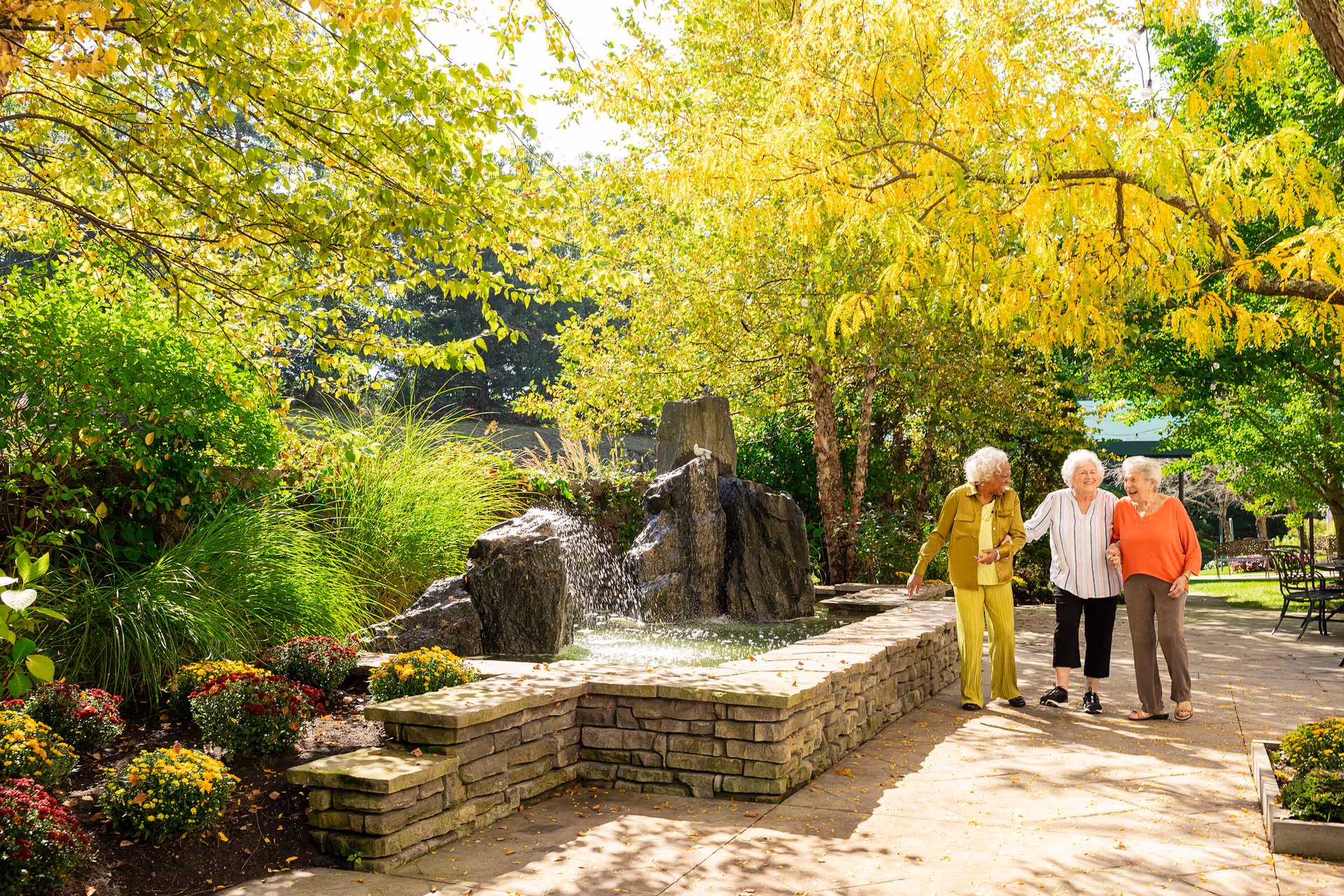 Three elderly women walking arm in arm along a sunny garden path beside a stone fountain and flowering shrubs.