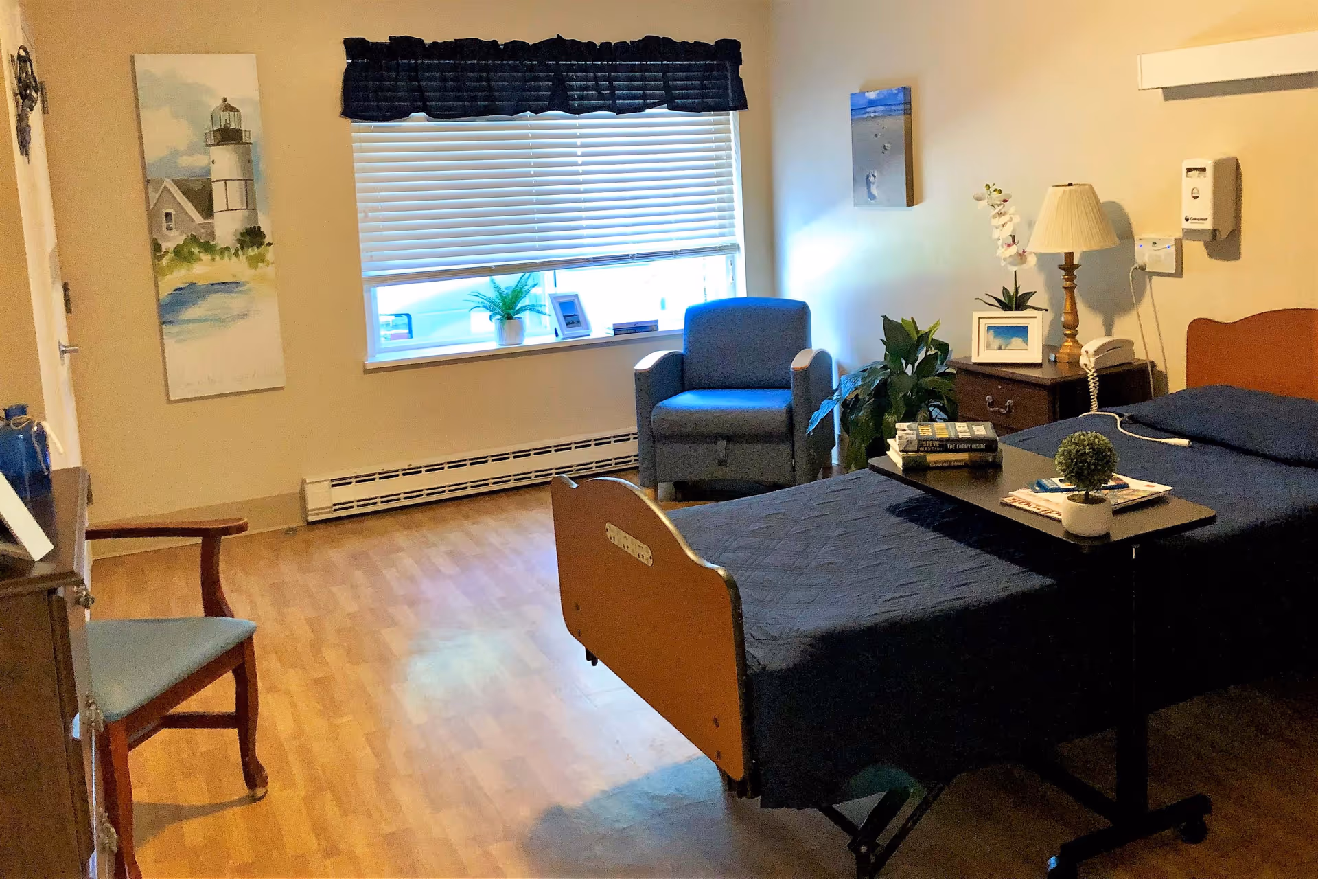 A well-lit room in a healthcare facility featuring a single hospital bed with a dark blue quilt, a small overbed table with books and a small plant, a wooden nightstand with a lamp, phone, and framed picture, a blue armchair near a window with blinds and a dark valance, a wooden chair, and wall decorations including a lighthouse painting and a beach-themed picture.