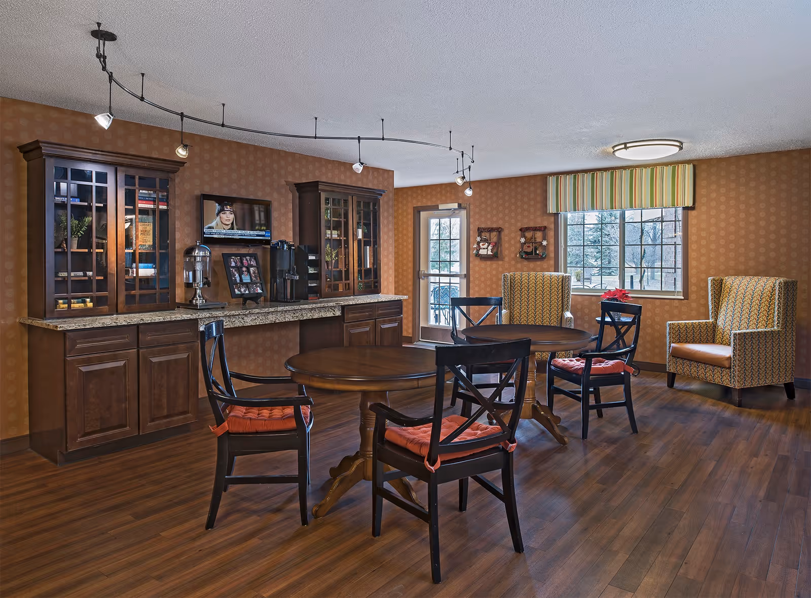 A cozy common area with two round wooden tables surrounded by black chairs with orange cushions. There are two patterned armchairs near a window with a striped valance. The walls have a warm patterned wallpaper, and there are two wooden cabinets with glass doors and a granite countertop along one wall. A small TV is mounted above the countertop, and a door leads outside.