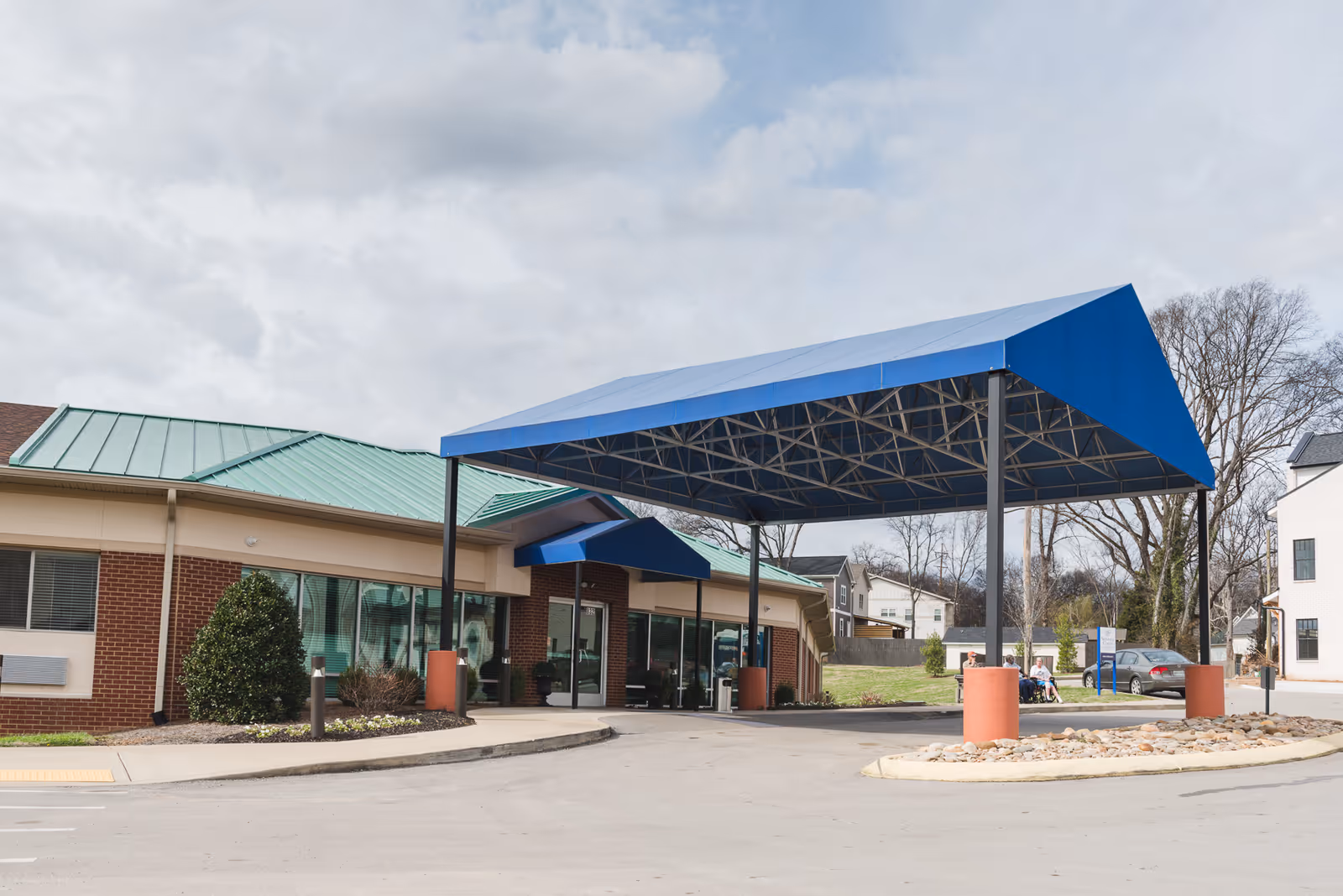 Exterior view of the Nashville Center for Rehabilitation & Healing showing the entrance with a large blue canopy supported by black metal poles. The building has a green metal roof and brick walls. There are some bushes and landscaping near the entrance, and a few people are visible sitting near the entrance area. The sky is cloudy.