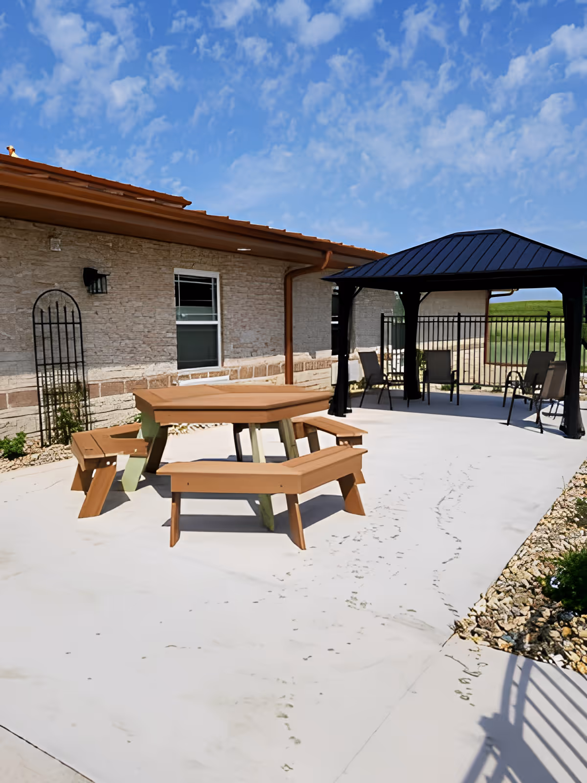Outdoor patio area at River Bend Retirement Community featuring a hexagonal wooden picnic table with attached benches on a concrete surface, a black metal gazebo with several chairs underneath, a stone building wall with a window, and a partly cloudy blue sky.