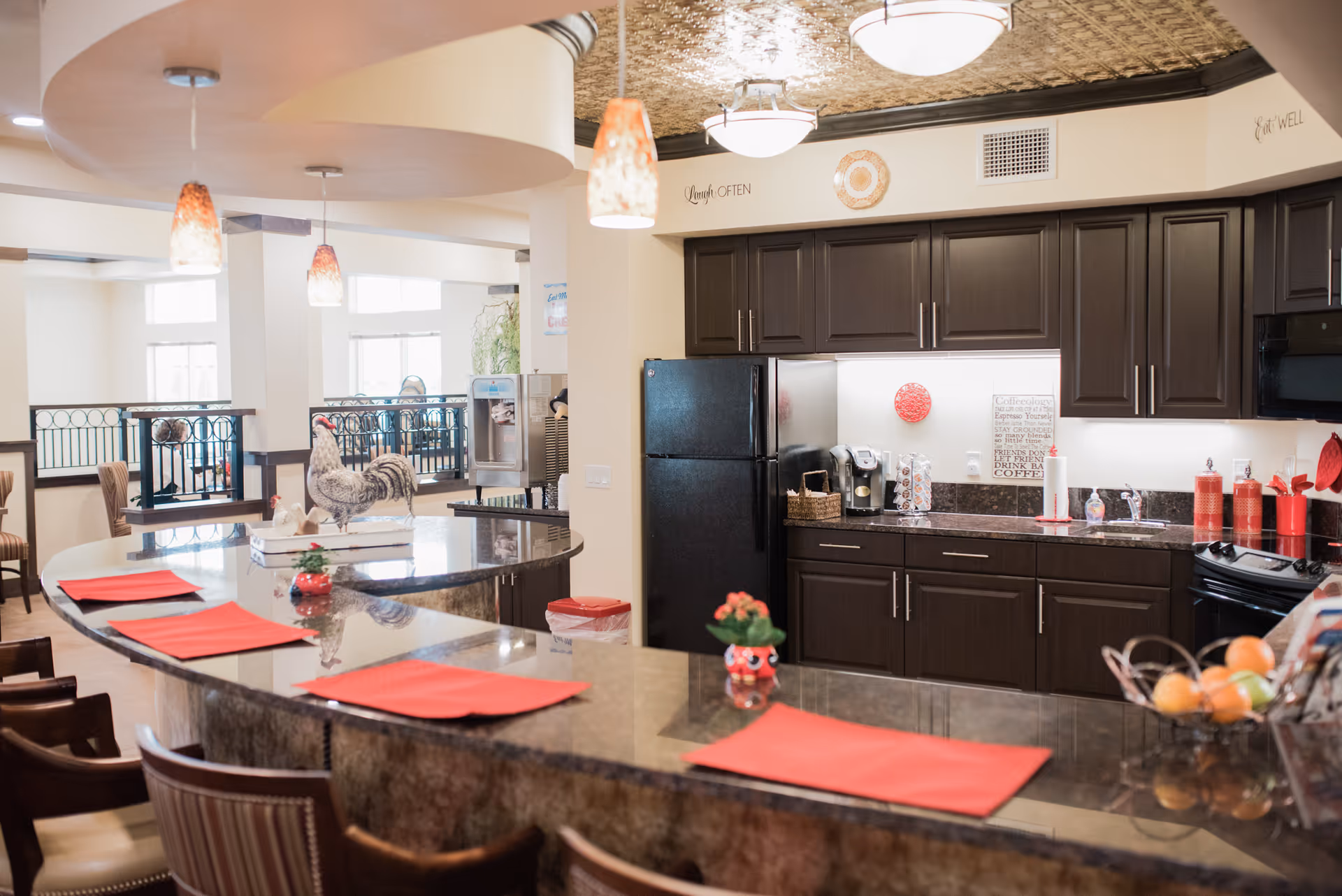 A modern kitchen area in a senior living facility with dark wood cabinets, a black refrigerator, and a curved granite countertop with red placemats. The kitchen features pendant lights, a coffee maker, and decorative items including rooster figurines and a fruit basket. There is a seating area with chairs along the counter and a water dispenser in the background.