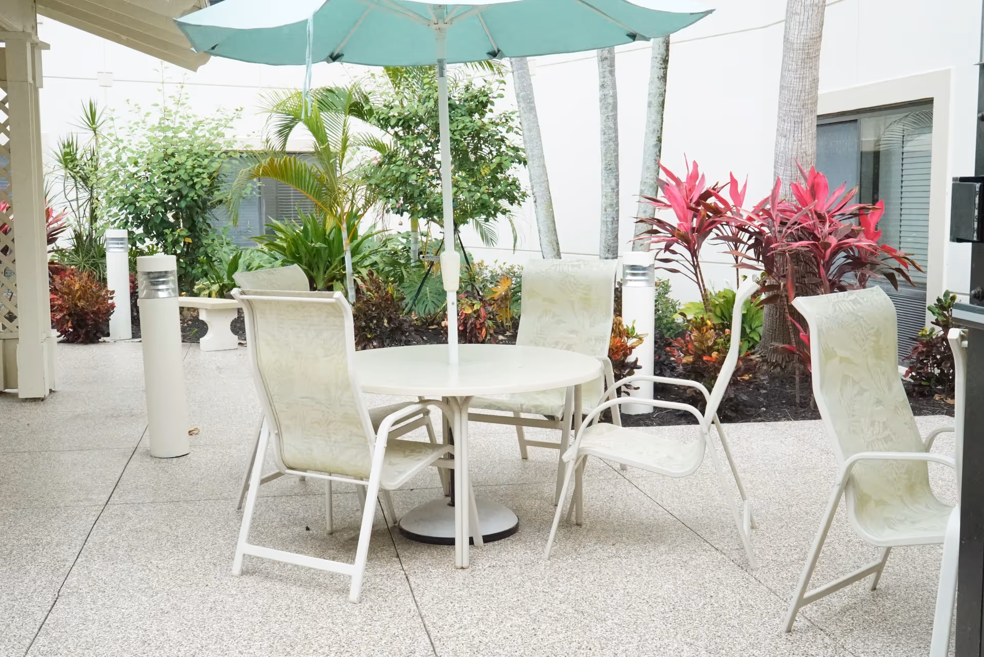 Outdoor patio area with a round white table and four white chairs with light-colored cushions. A large teal umbrella is positioned over the table. Surrounding the patio are various green plants and shrubs, including some with red leaves. The area is paved with light-colored stone tiles.