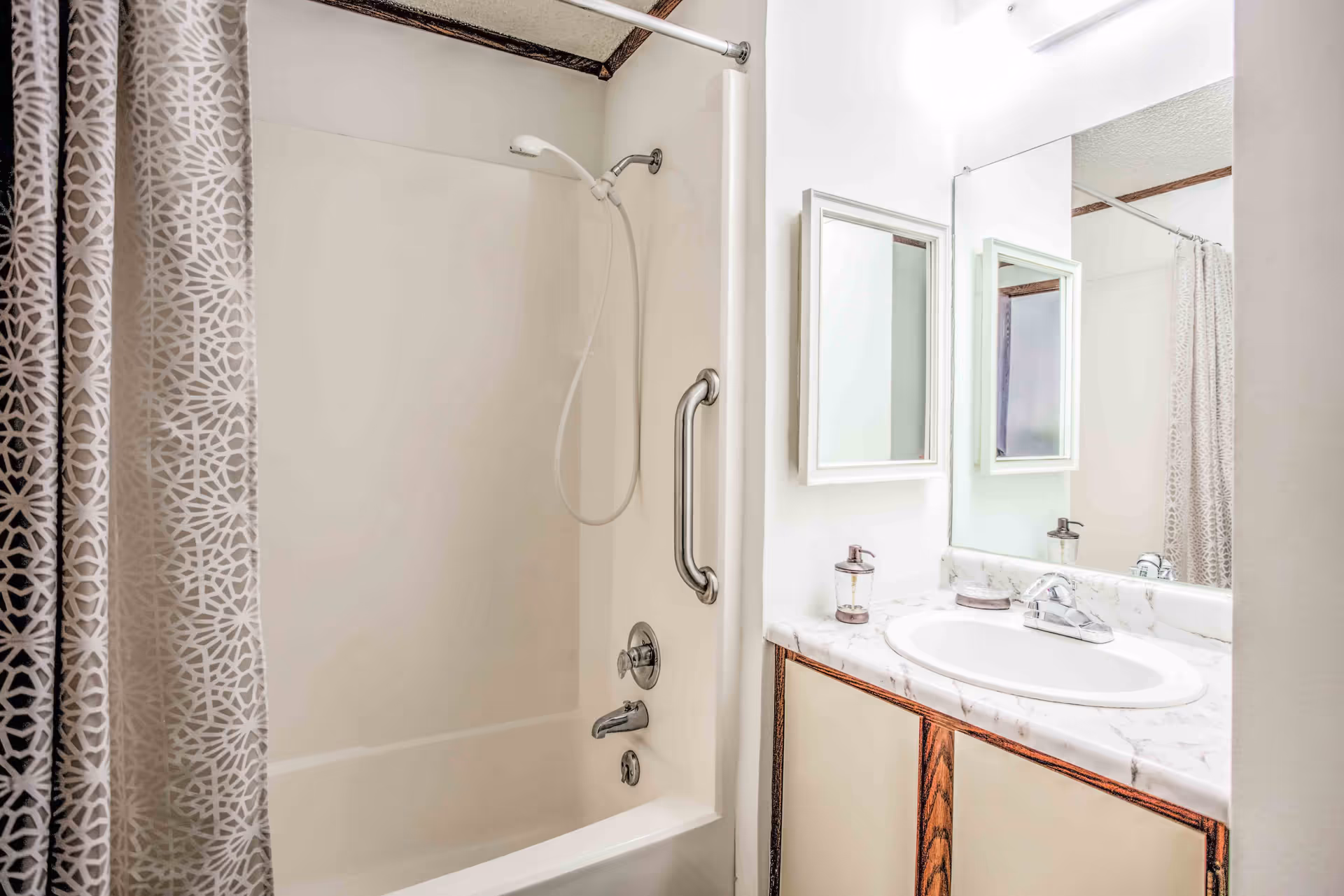 A bathroom featuring a white bathtub with a handheld showerhead and a patterned shower curtain. There is a metal grab bar on the bathtub wall. Next to the bathtub is a vanity with a marble countertop, a white sink, a soap dispenser, and a wall-mounted mirror above it.