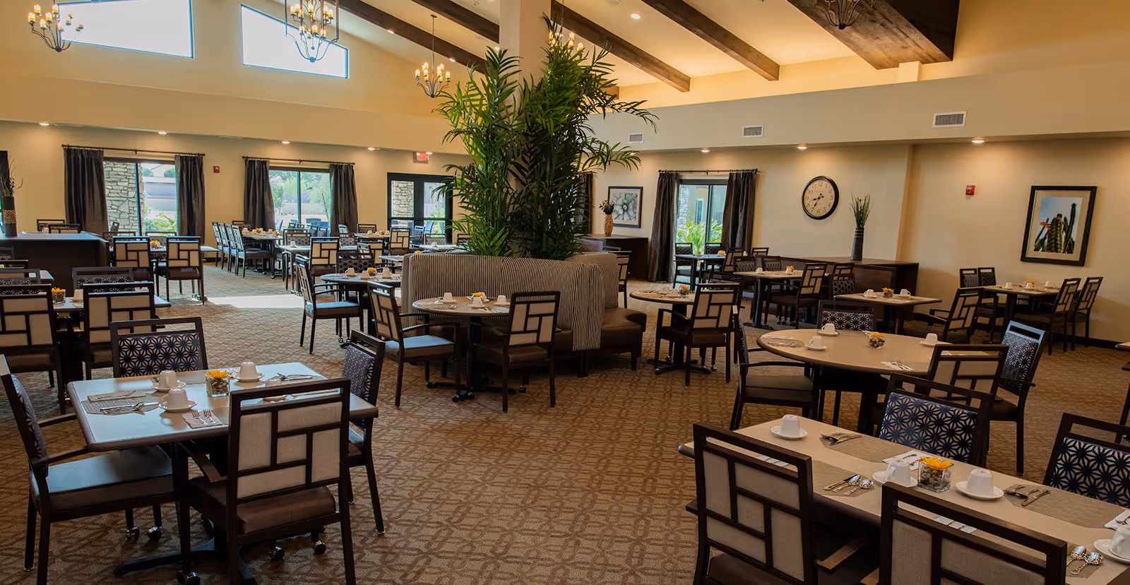 A spacious dining room with multiple tables and chairs arranged neatly. Each table is set with white cups, saucers, silverware, and small flower arrangements. The room features high ceilings with wooden beams, large windows with dark curtains, and decorative plants in the center. Wall decorations include a clock and framed artwork.