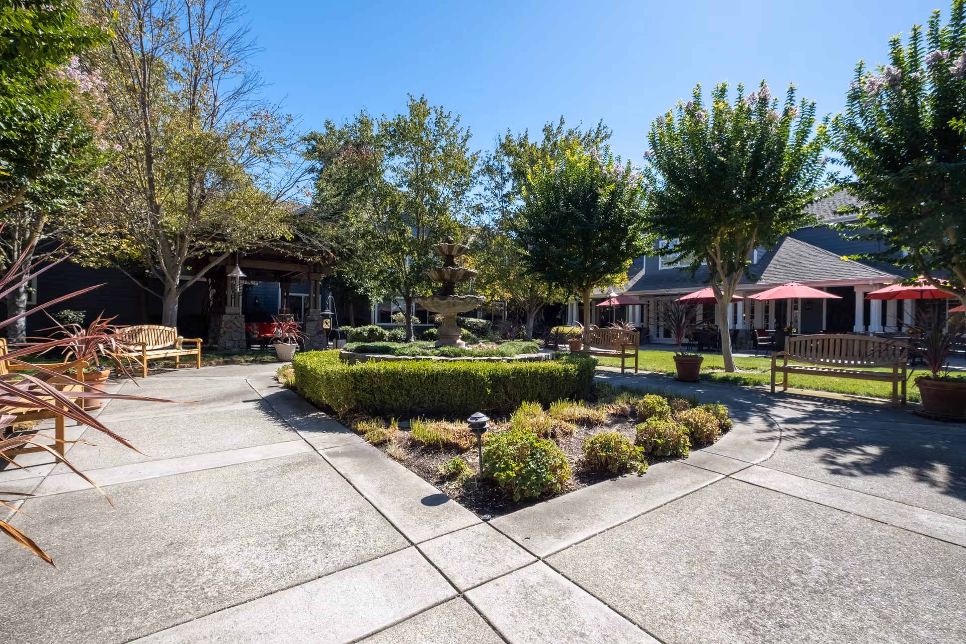 Sunny courtyard with a central fountain, benches, trees, potted plants and red patio umbrellas.