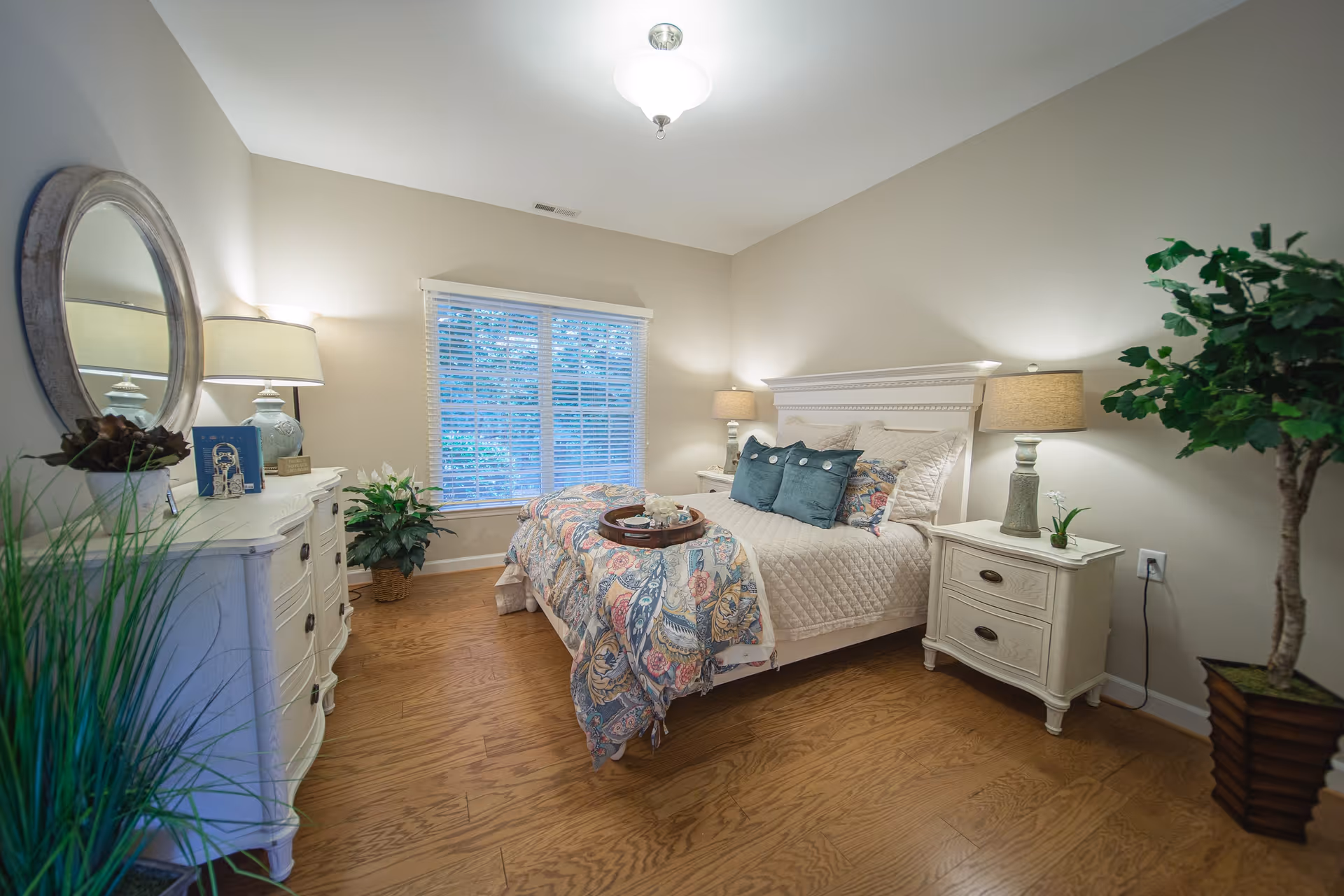 Bright, tastefully decorated bedroom with a bed, nightstands, dresser, plants, and a window with blinds.