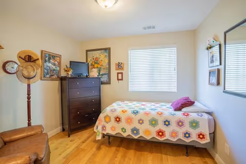 A cozy bedroom with a single bed covered by a colorful floral quilt. There is a dark wooden dresser with a small TV on top, and several framed pictures and paintings on the walls. A window with white blinds lets in natural light. A brown leather chair and a wooden hat stand with hats are also visible. The floor is light wood, and the walls are painted a soft beige.