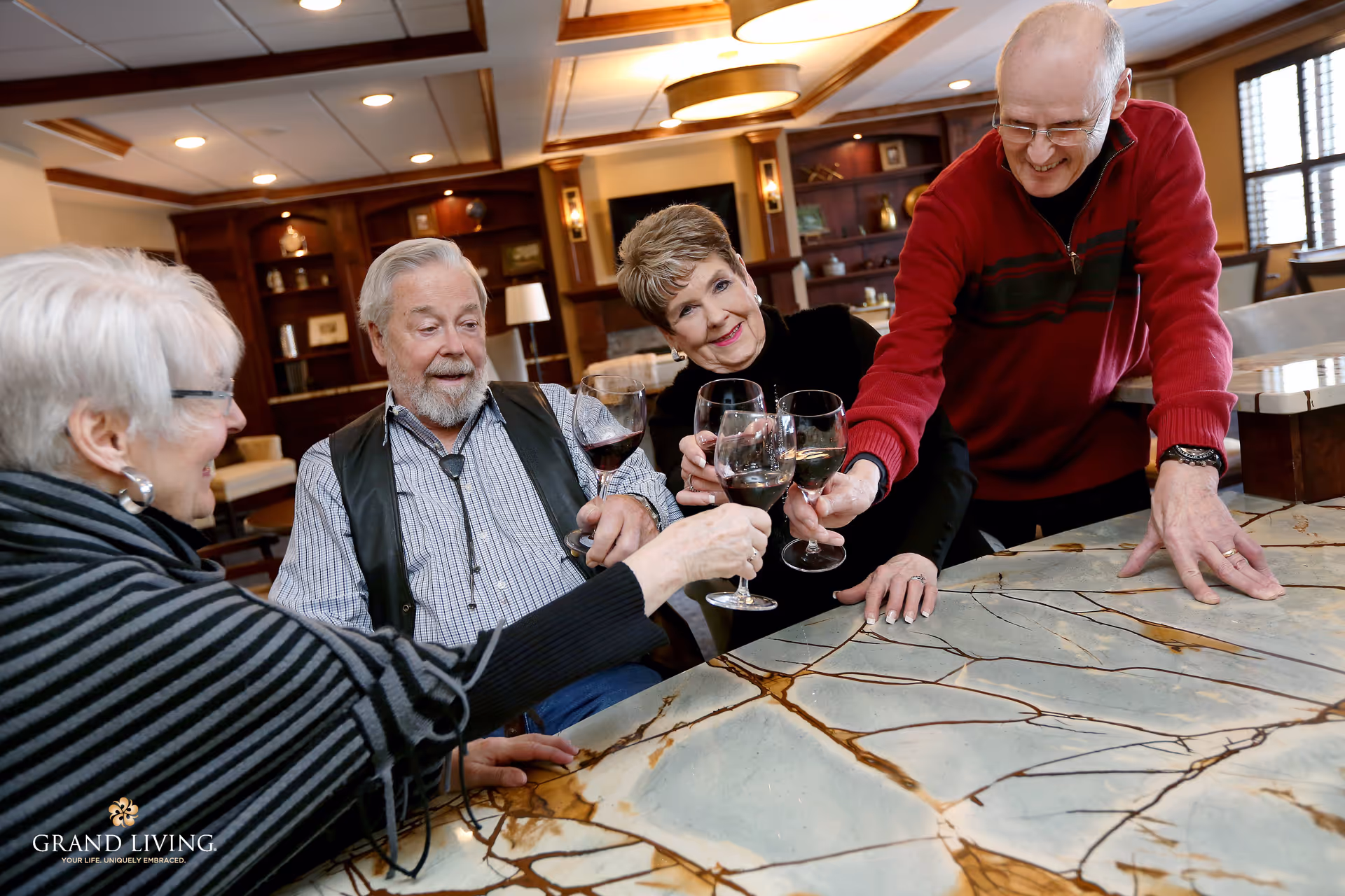 Four elderly people sitting around a marble table in a warmly lit room, smiling and clinking their wine glasses together in a toast. The background shows wooden shelves with decorative items and a cozy ambiance.