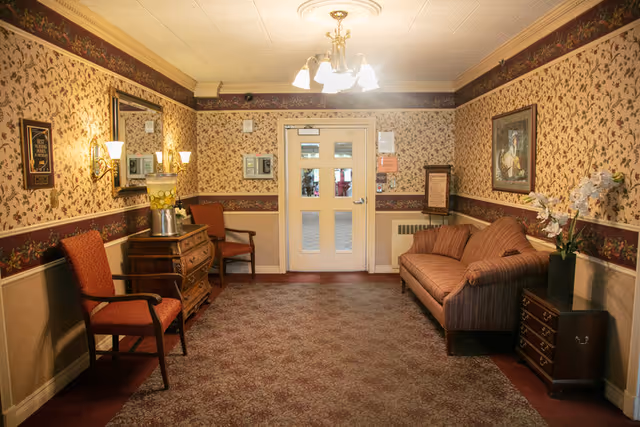 A cozy sitting area with floral wallpaper and a patterned carpet. The room features a striped sofa on the right with a small wooden side table holding a vase of white flowers. On the left, there are two red upholstered chairs and a wooden chest of drawers with a lamp on top. A large mirror hangs on the left wall above the chest. A chandelier with multiple lights hangs from the ceiling, and a door with glass panels is centered at the far end of the room.