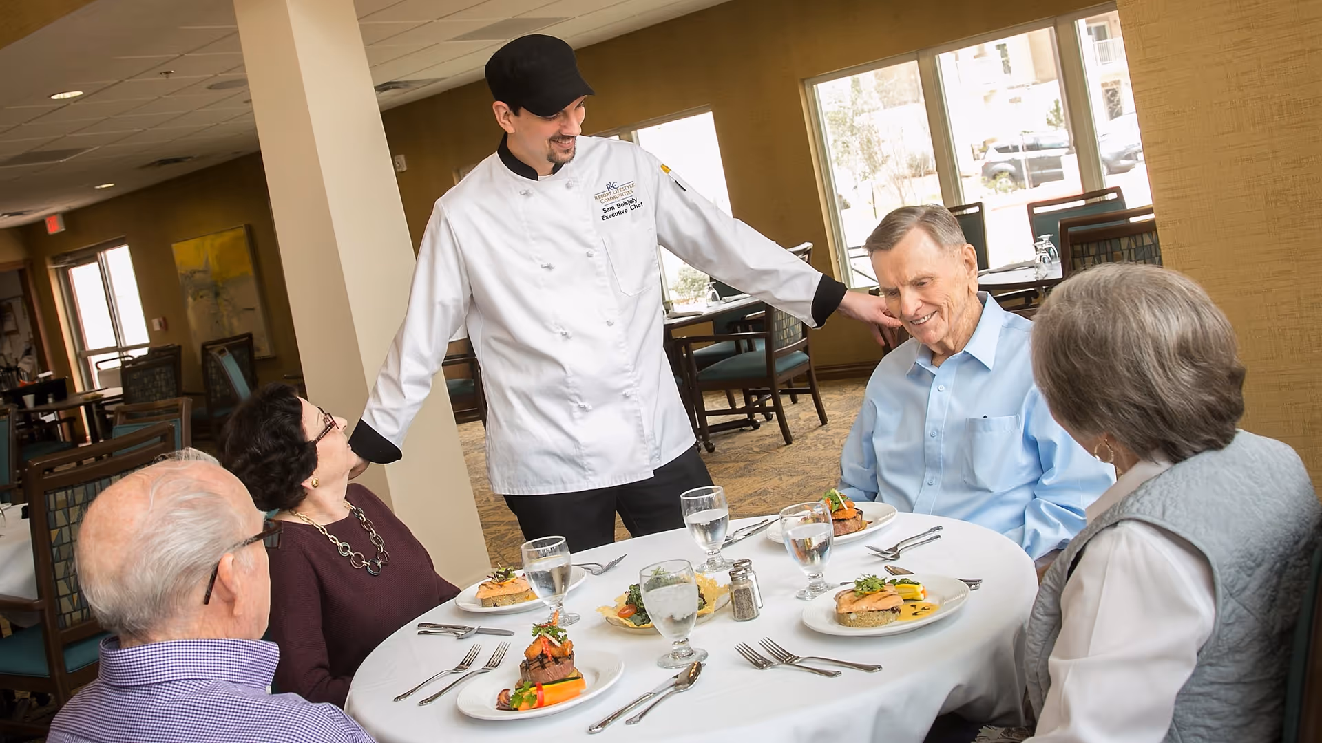 A chef talks with four elderly residents seated around a table set with plates and glasses in a dining room.