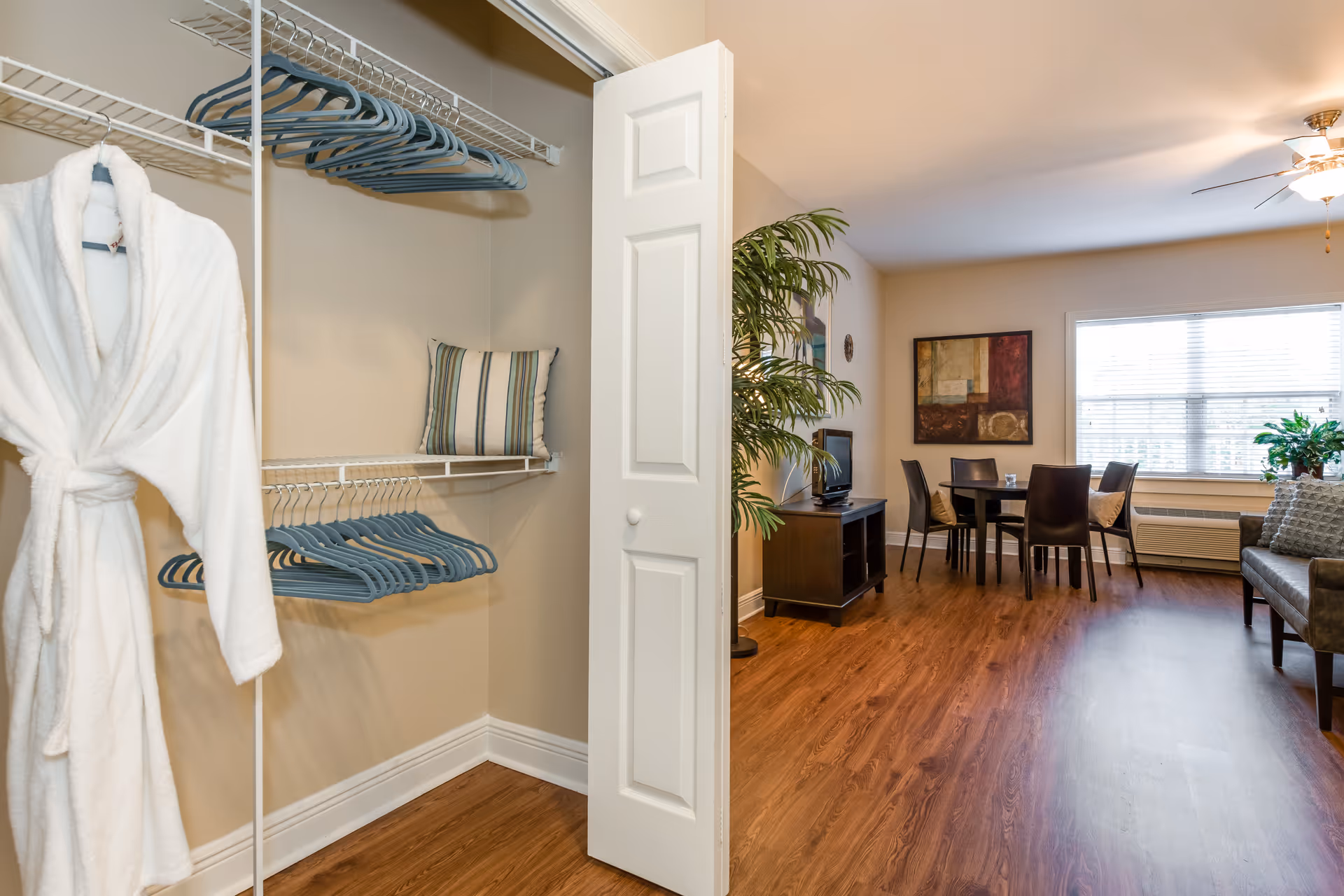 Open closet with hangers and a white robe opening into a furnished living and dining area with hardwood floors.