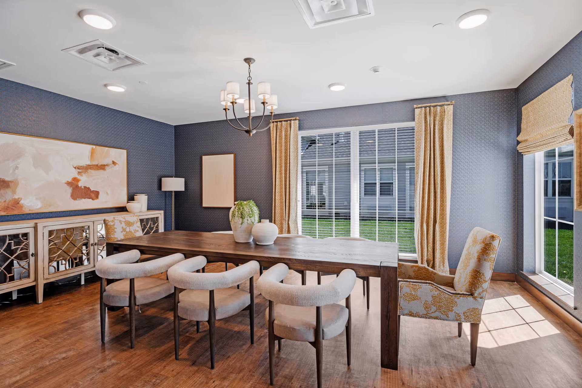 A bright dining room with a large wooden table surrounded by six upholstered chairs. The room features blue patterned walls, a wooden sideboard with decorative items, two large windows with beige curtains and blinds, and a chandelier hanging from the ceiling. Sunlight streams in through the windows, illuminating the space.