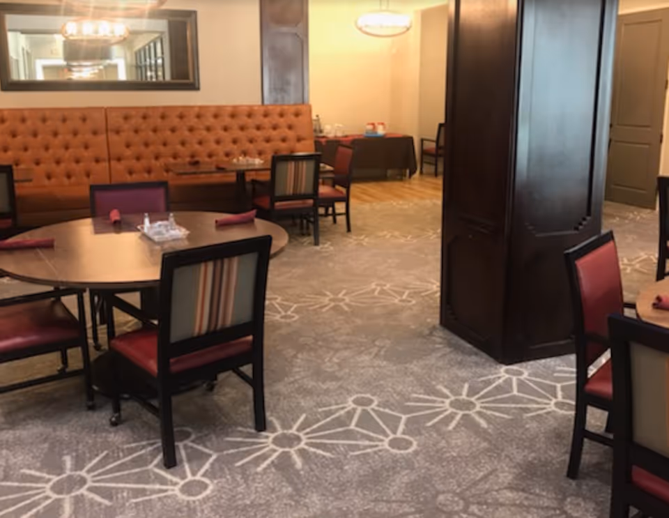 Dining room with round tables, upholstered chairs, a tufted banquette along the wall, and a central wooden column.
