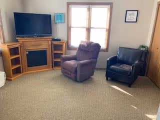A cozy living room with a large window letting in natural light. The room features a flat-screen TV on a wooden stand with built-in shelves, a purple recliner chair, a black leather armchair, and a beige carpeted floor. The walls are light-colored with minimal decorations.