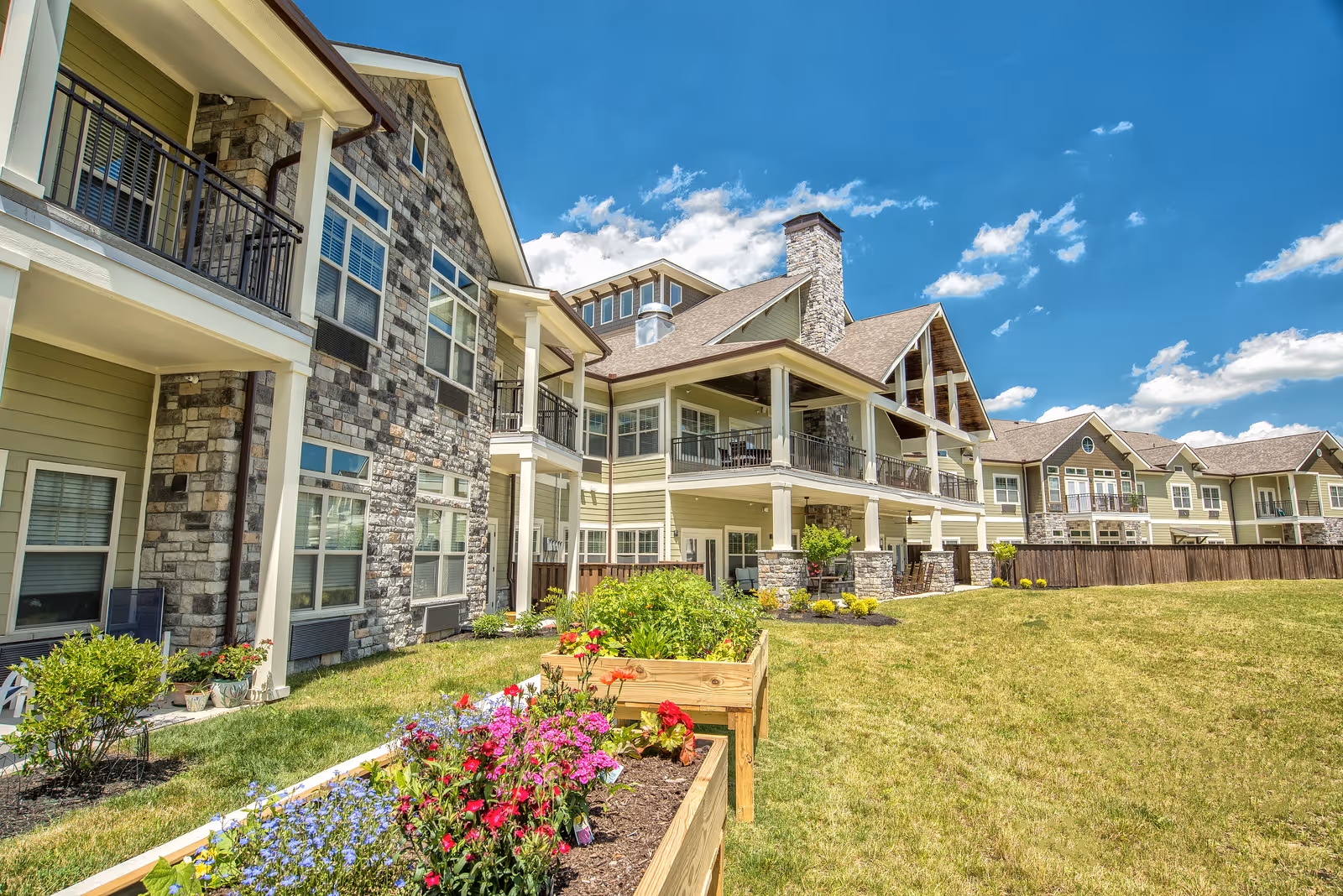 Exterior view of a multi-story senior living building with stone accents, covered balconies, a lawn, and raised flower beds.