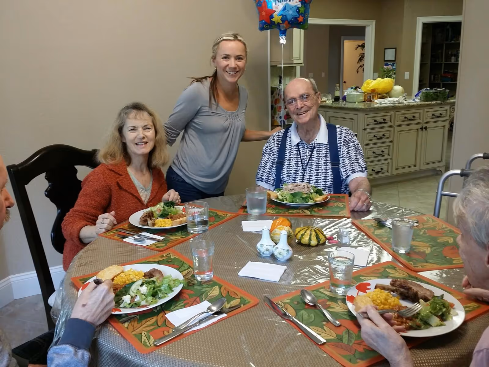 A group of elderly people and a caregiver gathered around a dining table enjoying a meal together in a senior living facility. The table is set with plates of food including salad, corn, and meat. The caregiver is standing and smiling behind two seated elderly individuals. The background shows a kitchen area with cabinets and countertops.
