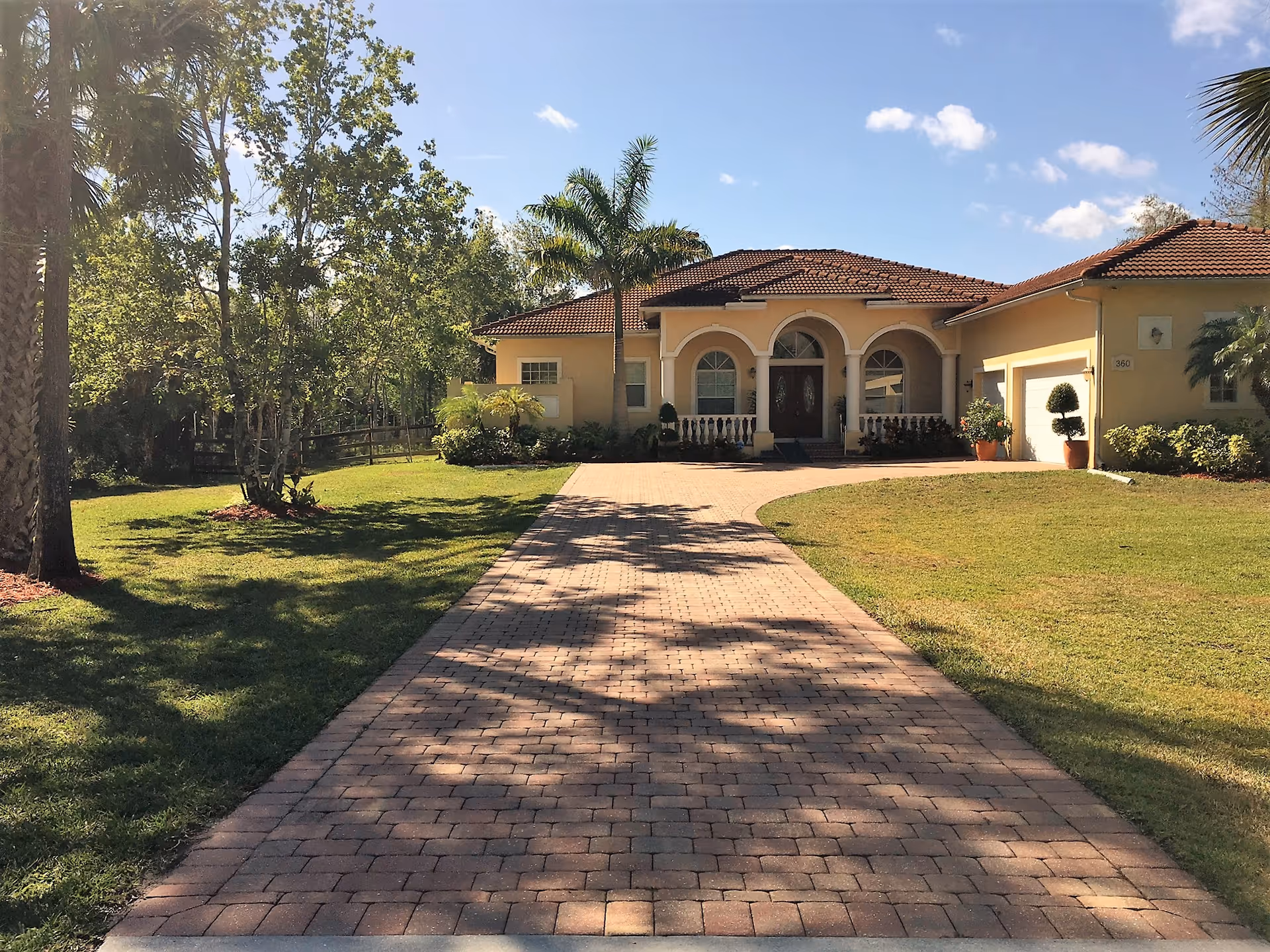 A single-story house with a tiled roof and arched front entrance, surrounded by a well-maintained lawn and palm trees. A paved driveway leads up to the front door, and the sky is clear with a few clouds.