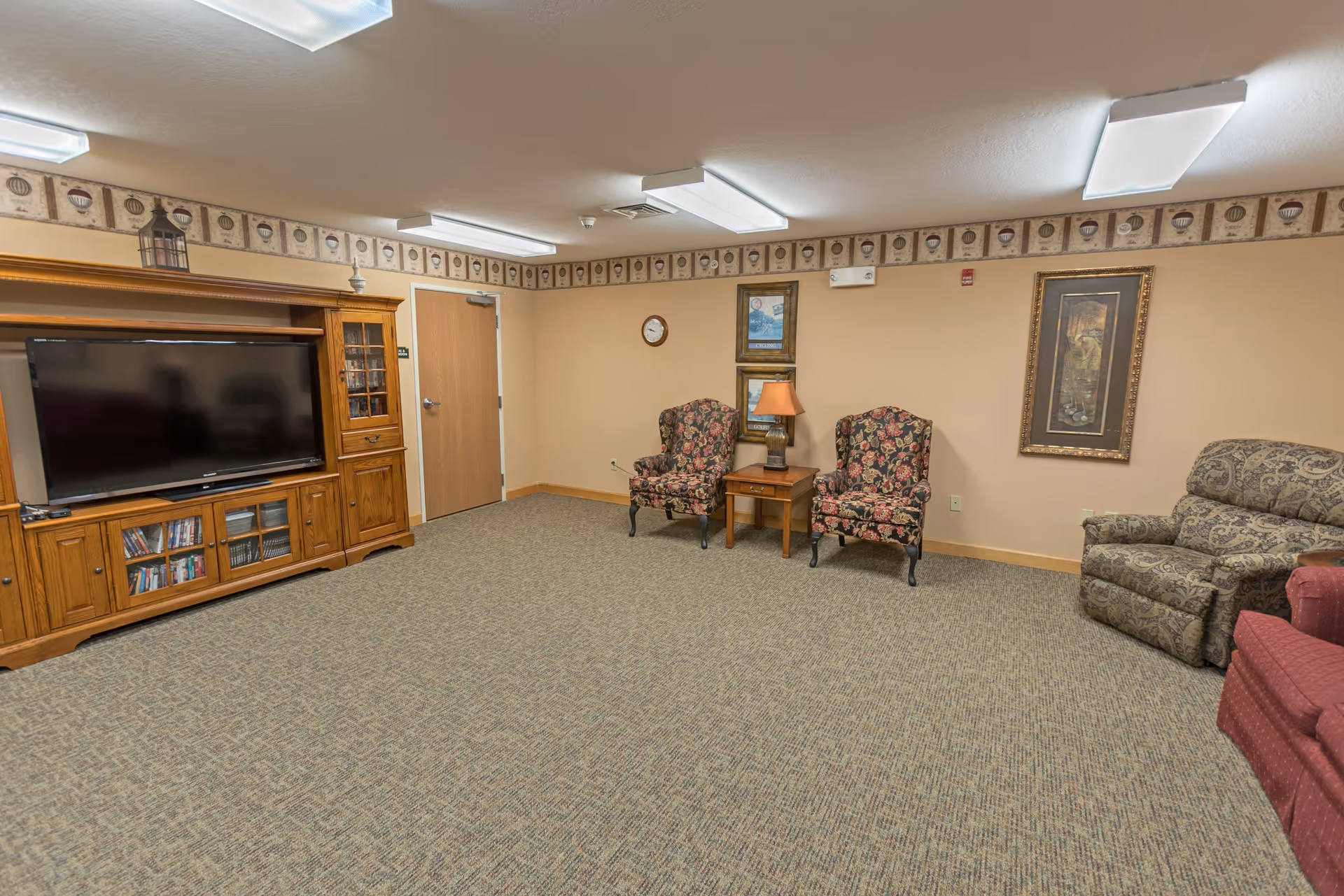 A common living area with a large wooden entertainment center holding a flat-screen TV and shelves with DVDs. The room has beige walls with a decorative border near the ceiling, carpeted floor, and fluorescent ceiling lights. There are two floral patterned armchairs with a wooden side table and lamp between them, a patterned recliner, and a red upholstered chair partially visible on the right.