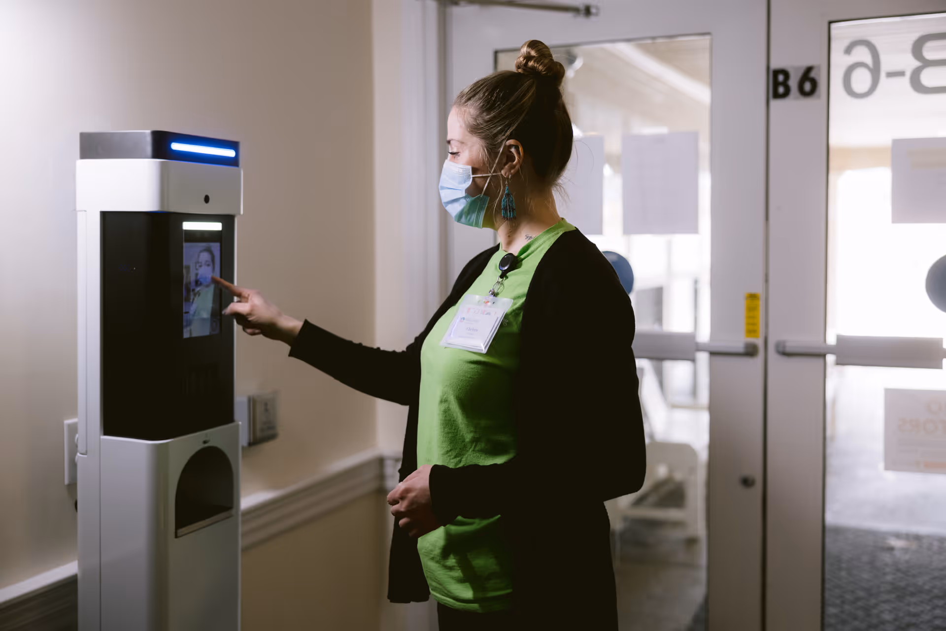 A woman wearing a green shirt, black cardigan, and a face mask is interacting with a touchscreen device mounted on a wall near an exit door inside a building.