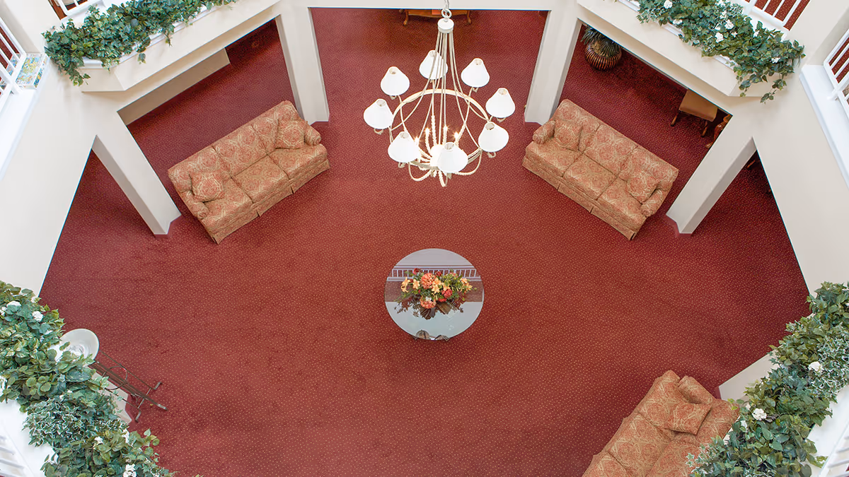 Overhead view of a spacious indoor lounge area with red carpet, three patterned sofas arranged around a circular glass table with a floral centerpiece, a large chandelier hanging from the ceiling, and green plants along the upper balcony railings.