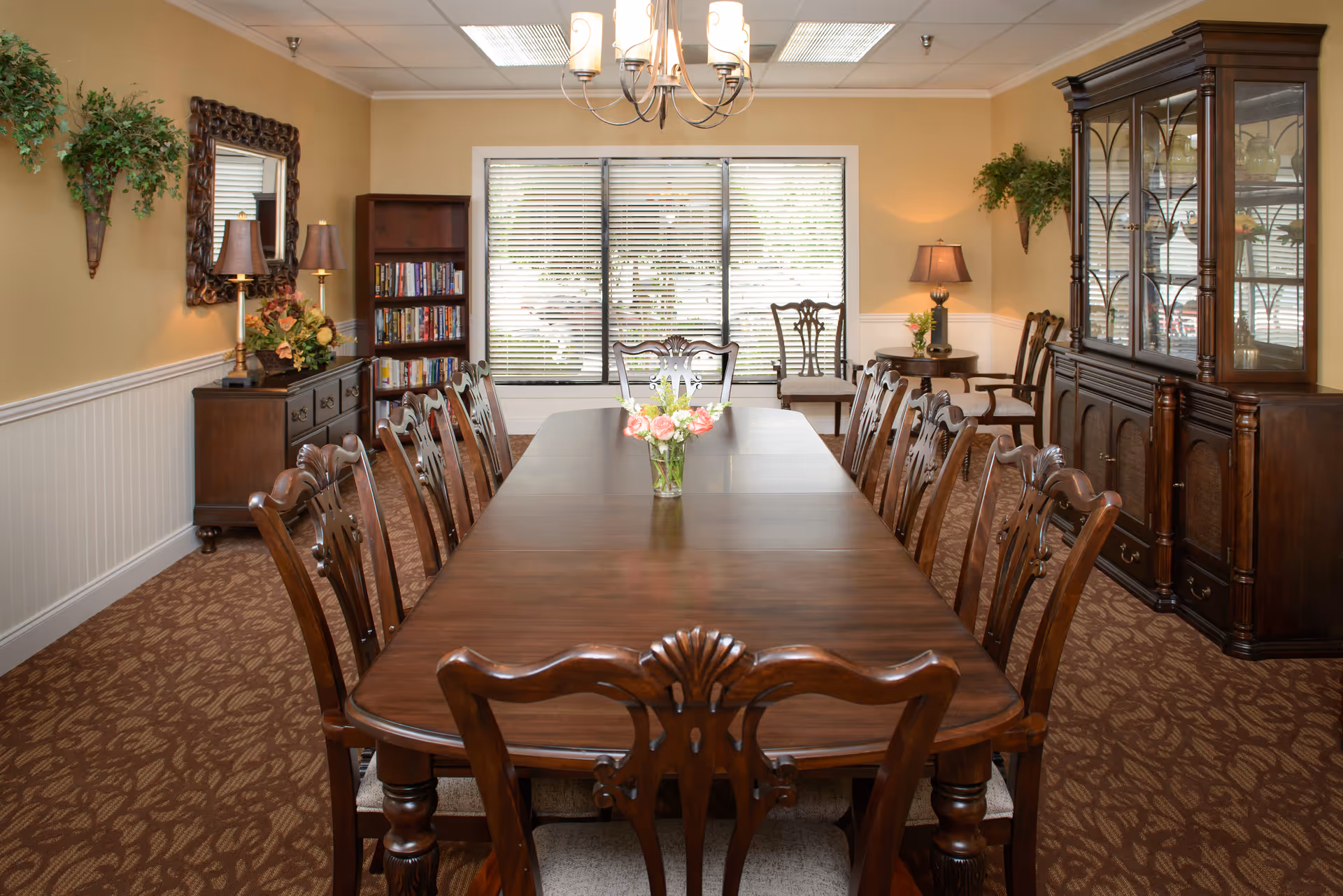 A formal dining room with a long wooden table surrounded by twelve matching wooden chairs. The table has a small vase with flowers in the center. The room features beige walls, a patterned carpet, a chandelier overhead, a large window with blinds, a wooden china cabinet, a sideboard with lamps and floral arrangements, a bookshelf, and some green plants on the walls.