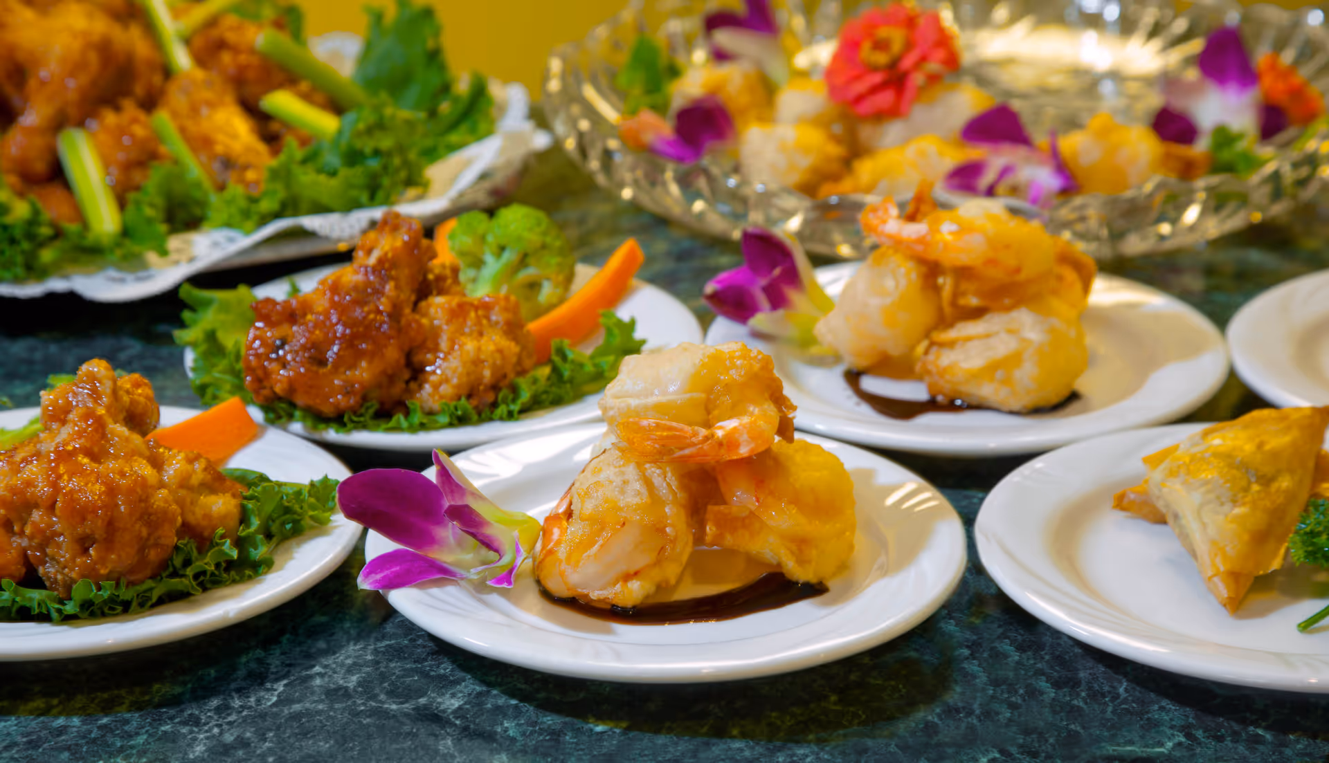 Several white plates with assorted appetizers including fried shrimp, fried chicken pieces, and a pastry, garnished with leafy greens, carrot sticks, broccoli, and purple edible flowers, arranged on a dark green marble countertop.