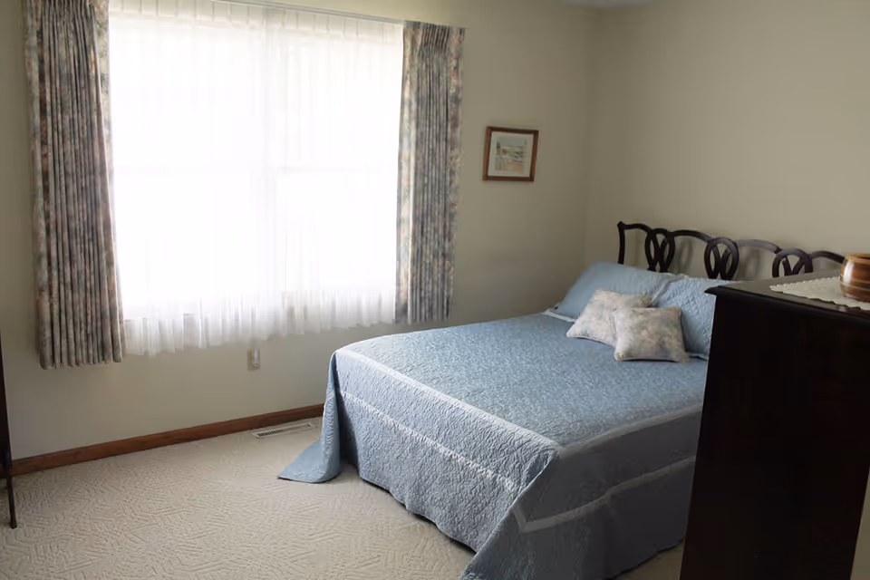 A bedroom with a bed covered in a light blue quilt and two matching pillows. There is a window with sheer white curtains and floral drapes letting in natural light. A framed picture hangs on the wall, and a dark wooden dresser is partially visible on the right side.