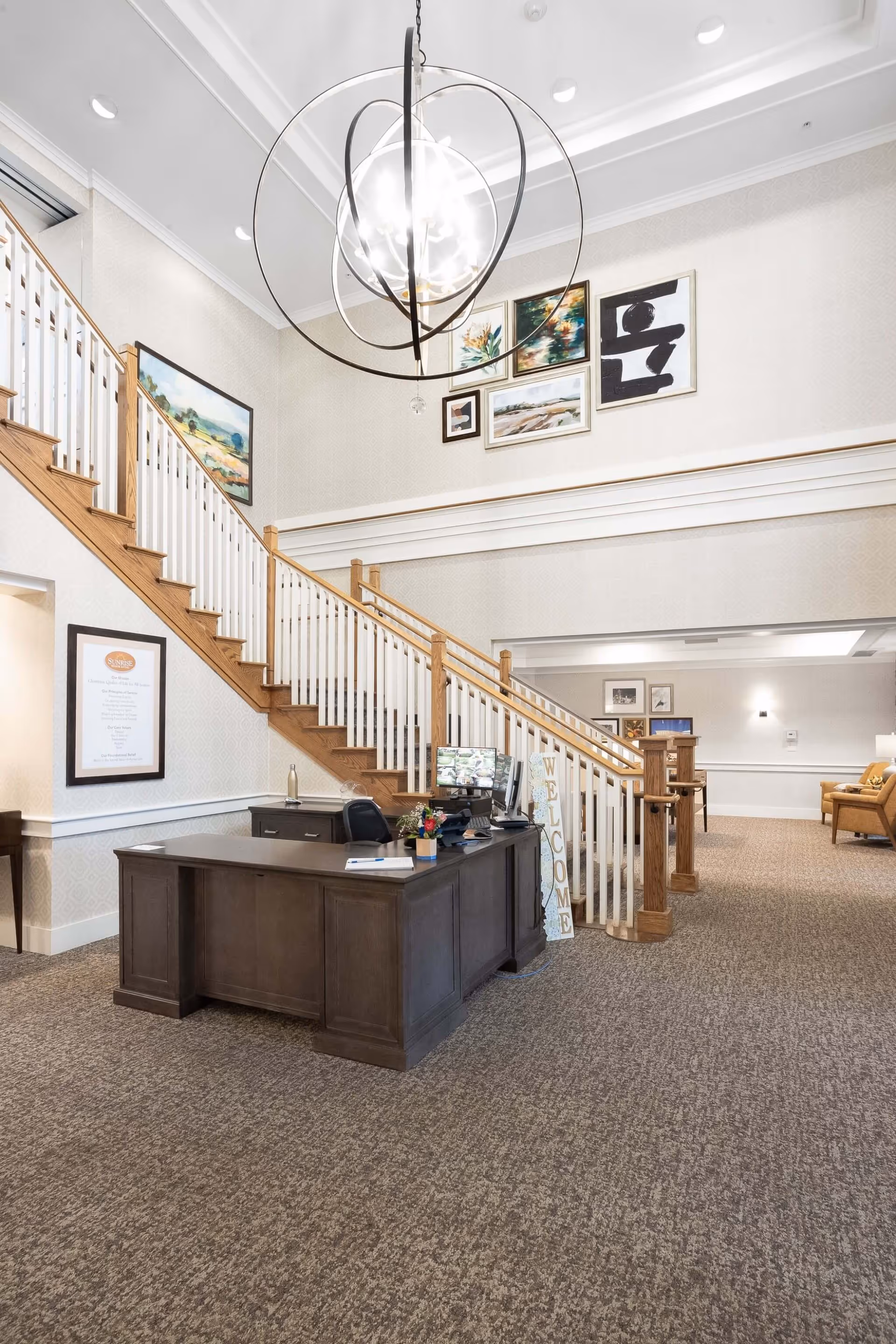 Interior view of a senior living facility lobby with a wooden reception desk, a computer monitor, and a welcome sign next to a staircase with wooden handrails and white balusters. The walls are decorated with framed artwork, and a large modern chandelier hangs from the ceiling. The carpet is a neutral tone, and there is a seating area with chairs visible in the background.