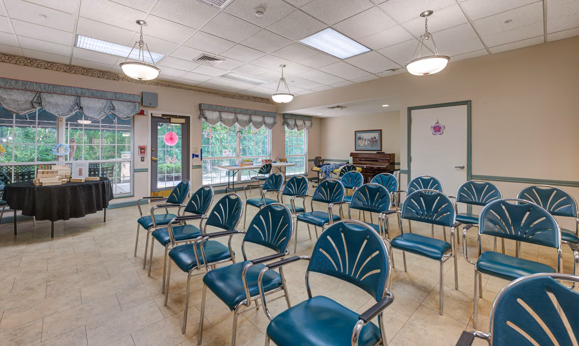 A community room with multiple rows of teal chairs with metal frames arranged facing forward. The room has large windows with valances, a door leading outside, a piano in the corner, and tables along the windows with various items on them. The walls are beige with green trim, and the ceiling has fluorescent lights and hanging light fixtures.